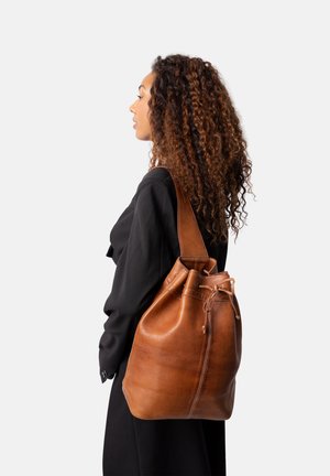 Sac en cuir marron de type bucket avec un cordon de serrage, présentant une texture lisse et une seule sangle, porté par une personne vêtue d'une chemise noire.