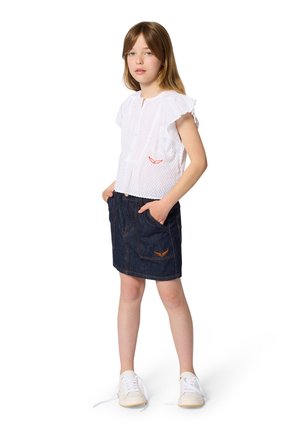 Jeune fille debout, les mains dans les poches, portant un chemisier blanc texturé à manches courtes, une jupe en denim foncé et des baskets blanches aux lacets dénoués.