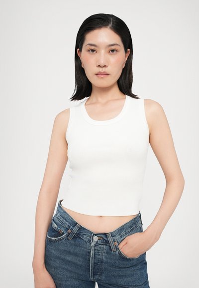 Un débardeur blanc à côtes sans manches avec un col rond, associé à un jean en denim bleu foncé taille haute avec fermeture à bouton et poches.