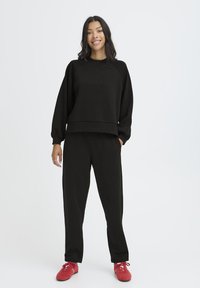 Melnā sporta krekls ar garām, pūkainām piedurknēm un apaļu kakla izgriezumu, apvienots ar atbilstošām melnām sporta biksēm, kurām ir sānu kabatas un brīvs piegriezums.