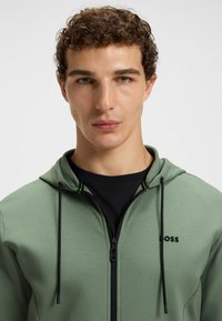 Hooded zip jacket in light green fabric with black accents. Features a smooth texture, zipper closure, and a subtle logo on the chest.