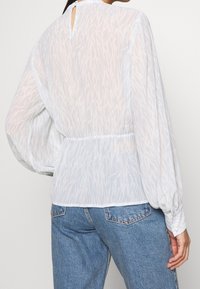 Femme portant une blouse blanche transparente à manches longues avec un motif ondulé subtil et un jean en denim bleu, vue de dos.