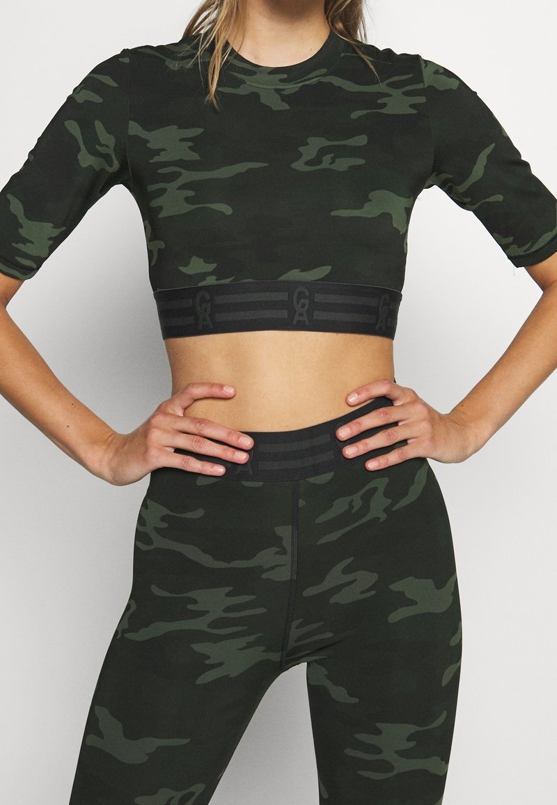 Lyhyet hihnat, tummanvihreä camo-kuvioinen crop-top ja legginsit, joissa on musta logolla varustettu elastinen vyö. Venytettävä kangas, tiukasti istuva malli.
