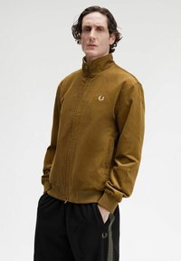 Uomo con le mani in tasca che indossa una giacca marrone in corduroy con zip e pantaloni neri con una striscia a motivi su uno sfondo chiaro e semplice.