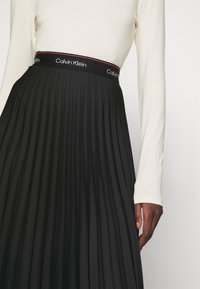 Gonna nera plissettata con vita elasticizzata che presenta il logo "Calvin Klein" rosso e bianco. La parte superiore è una camicia a maniche lunghe di colore chiaro.