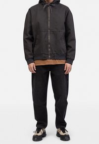 Sort hættetrøje med lynlås foran, to lommer og et beige inderlag, kombineret med mørke jeans og beige sportssko.