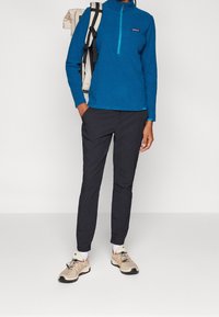 Modrý fleecový pulovr s polovičním zipem, černé kalhoty a béžové tenisky. Má texturovanou tkaninu a logo na hrudi.