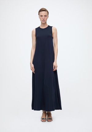 Γυναίκα στέκεται προς τα εμπρός φορώντας ένα αμάνικο μακρύ φόρεμα σε ναυτικό μπλε και ασημένια σανδάλια με ανοιχτά δάχτυλα, μπροστά σε ένα απλό λευκό φόντο.