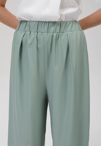 Pantalon vert clair à jambes larges avec une taille élastique et des plis, fabriqué dans un tissu lisse et texturé. Aucun détail métallique ou motif visible.