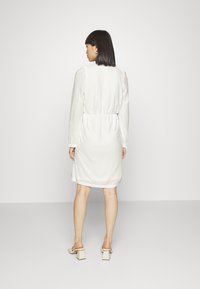 Vestido branco com padrão texturizado, cintura franzida e mangas longas. Modelo usa mules de salto bloco, exibindo um design simples e descontraído.