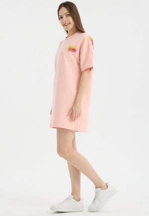 Jeune femme portant une robe t-shirt oversize rose clair avec un motif coloré et des baskets blanches, debout de côté, souriante.
