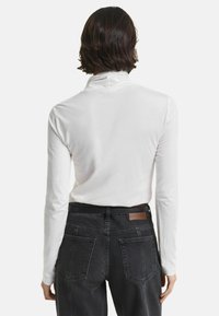 Persoon met kort bruin haar, gekleed in een witte lange mouwtop en zwarte hoge taillejeans, gezien vanachter tegen een effen achtergrond.