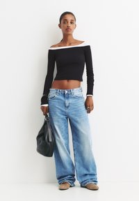 Schwarzes Off-Shoulder-Oberteil mit langen Ärmeln, kombiniert mit hellblauen weit geschnittenen Jeans. Accessoires sind eine schwarze Tasche und beigen Slipper.