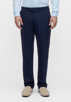 Homme portant un pantalon de costume bleu marine à fines rayures, une chemise bleu clair à manches longues et des mocassins beiges, debout devant un fond blanc.