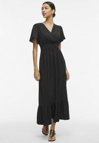 Must mustriga musta kleit lühikeste lennukite varrukatega, smockitud vöökohaga ja laieneva volangiga äärega. Kandes pruunide sandaalide ja sileda kangastekstuuri.