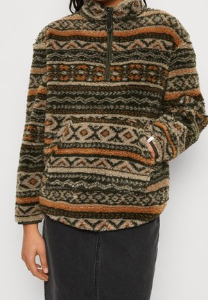 Personne portant un pull doux à motifs marron, vert et orange avec une poche avant et une fermeture éclair au quart, associé à une jupe foncée.