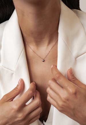 Nahaufnahme einer Frau, die ein weißes Blazer justiert und eine zarte Halskette mit einem kleinen herzförmigen Anhänger trägt.