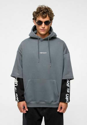 Grijze oversized hoodie met ribbelstructuur, voorzak en de tekst "IMPACT" in het wit gedrukt. Lange zwarte mouwen met witte tekst. Casual ontwerp.