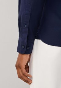 Camicia blu navy a maniche lunghe con polsino abbottonato e trama liscia. Un primo piano mostra il dettaglio del polsino e una mano che tiene il lato della camicia.
