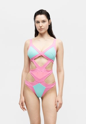 Tânără cu păr ud, purtând un costum de baie intreg roz și albastru, cu decupaje, stând pe un fundal alb simplu.