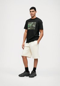 T-shirt noir à manches courtes avec un graphique vibrant de crocodile et le texte « LACOSTE ». Assorti à un short crème et des chaussures de sport noires.