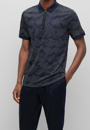 Homme portant un polo bleu marine à manches courtes avec col zippé et motif géométrique à pois, associé à un pantalon bleu marine foncé.