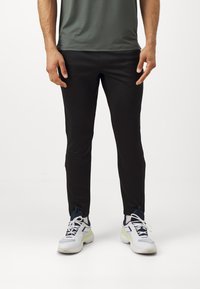 Fekete sportos nadrág, tapered fazonnal és sima textúrával. Fehér és sötétkék sportcipővel párosítva, fényvisszaverő részletekkel.