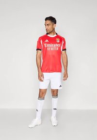 Červený futbalový dres Adidas s čiernymi detailmi a bielym nápisom, doplnený o biele šortky a kolienkové ponožky s čiernymi pruhmi.