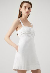 Vestido de lino blanco con un bodice ajustado que presenta pliegues horizontales, tirantes estrechos y bolsillos laterales. Silueta en A con un dobladillo limpio.