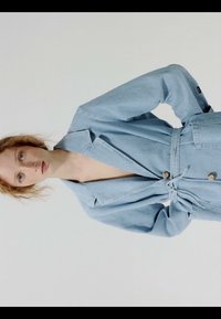 Mujer con cabello rizado y rojo que lleva un abrigo azul claro con cinturón, de pie con las manos en las caderas frente a un fondo blanco liso.