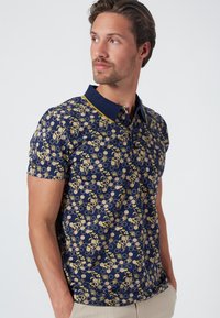 Korte mouwen polo shirt in donkerblauw met een floraal en hexagonaal patroon in geel en bruin. Beschikt over een contrasterende marineblauwe kraag en knoopsluiting.