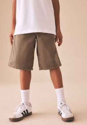 Kind draagt bruine kniebroek, witte sokken en witte Adidas sneakers met zwarte strepen, staand op een beige vloer.