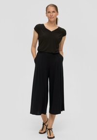 Zwarte top met een V-hals en gathered schouderdetails, gecombineerd met brede zwarte broek met zakken en een vloeiende pasvorm. Platte sandalen.