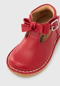 Κόκκινο δερμάτινο παπούτσι για νήπια με ραμμένη σόλα, λουράκι σε σχήμα Τ, διακοσμητικό φιόγκο και ασημένια αγκράφα κλεισίματος σε λευκό φόντο.