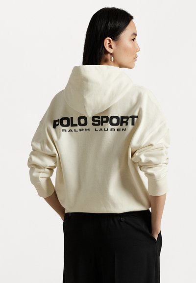 Kremfarget hettegenser med en stor svart "POLO SPORT RALPH LAUREN"-logo på ryggen. Har hette og ribbede mansjetter, brukt over svarte bukser.