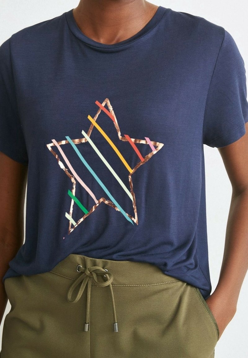 Personne portant un t-shirt bleu marine avec une grande étoile soulignée en cuivre et remplie de rayures diagonales colorées, associée à un pantalon vert olive.