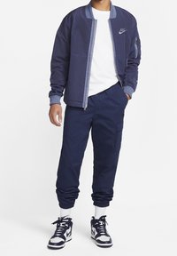 Marinblå bomberjacka med dragkedja fram, ribbade ärmar och Nike-logotyp; kombinerad med marinblå cargo-byxor och högskor. Vit t-shirt under.