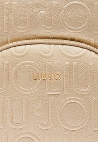 Borsa in pelle texturizzata beige chiaro con design del logo impresso. Un accento di hardware in tonalità oro è visibile vicino alla cerniera.
