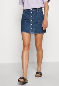 Jupe en jean à coupe ajustée, de couleur bleu foncé, avec une fermeture à boutons sur le devant, des passants pour ceinture, et un ourlet effiloché. Assortie à des sandales noires.