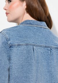 Chaqueta de mezclilla azul claro con un yugo trasero, detalles de costura mínimos y un corte relajado. Textura suave, cuello estándar y sin cierres visibles.