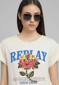 Maglietta beige con scritta "REPLAY" blu e grafica di una rosa rosa. Il modello indossa occhiali da sole con montatura argento. Tessuto liscio, design casual.
