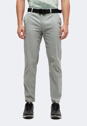 Homme portant un pantalon gris clair avec une ceinture noire et des chaussures noires, debout, les mains relâchées le long du corps sur un fond uni.