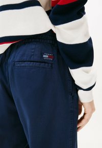 Granatowe bawełniane spodnie z tylną kieszenią, z czerwoną i białą metką "TOMMY JEANS". Górna część przedstawia sweter w paski w kolorze czerwonym, białym i granatowym.