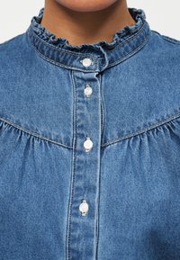 Nærbilde av en person som har på seg en blå denimskjorte med perleknapper og en rysjekrage.