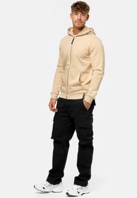 Beige zip-up hoodie med snoretræk i hætten, med et logo på fronten. Parret med sorte cargo-bukser og hvide sneakers.