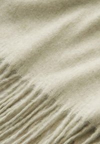 Tissu en laine beige doux avec des franges le long d'un bord, mettant en valeur la texture et les fibres.
