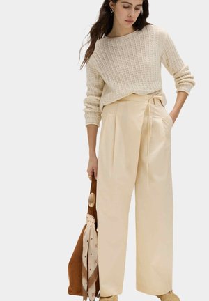 Donna che indossa un maglione crema in maglia e pantaloni beige a vita alta a gamba larga, che tiene una borsa marrone con sciarpa fantasia su sfondo neutro.