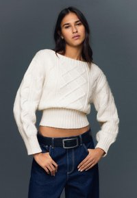 Fehér, kábelmintás, rövid felső hosszú ujjakkal, bordázott szegéllyel. Sötétkék, magas derekú farmerral és fekete övvel párosítva.