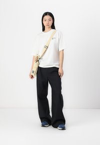 T-shirt blanc avec un petit logo, pantalon noir à jams larges et baskets bleues. Un sac en bandoulière beige est porté en travers de l'épaule.
