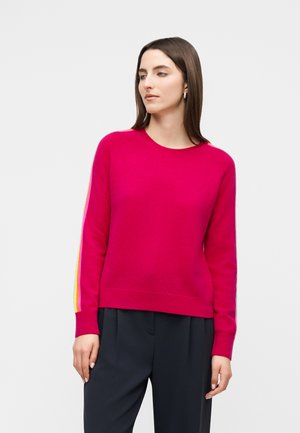 Vrouw draagt een felroze trui met gele en witte strepen op de mouwen, gecombineerd met een zwarte broek, staand tegen een effen achtergrond.